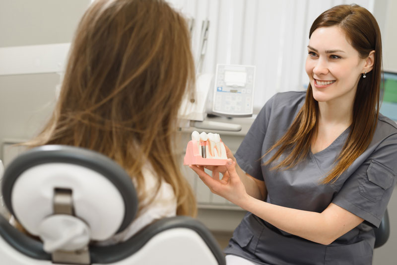 a dental assistant showing a patient a dental implant model so she can explain to her why she needs to be treated with dental implants.