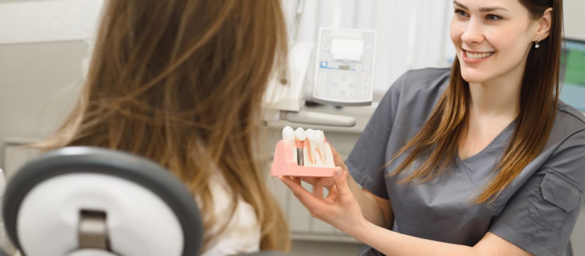 a dental assistant showing a patient a dental implant model so she can explain to her why she needs to be treated with dental implants.